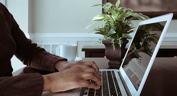 Person typing on a laptop at a desk with a coffee mug and houseplant, reading real estate articles